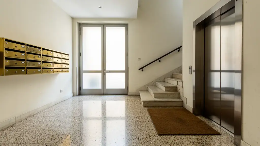 Androne di un condominio con cassette postali in ottone, portone a vetri, pavimento in graniglia, scale in marmo e ascensore